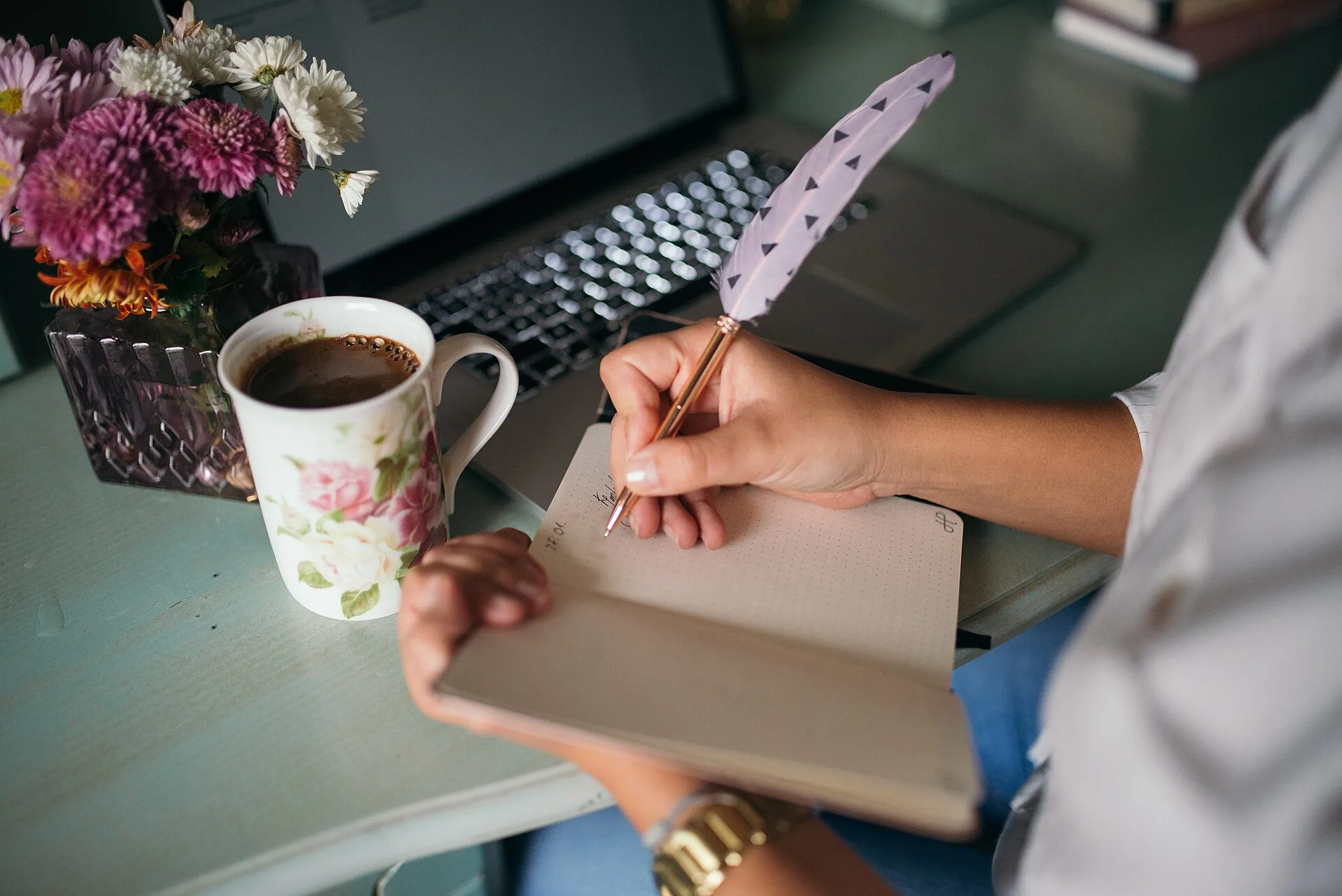 A person is writing in a blank notebook with a feather pen at a desk. A cup of coffee sits nearby, alongside a bouquet of colorful flowers, creating a cozy workspace vibe. Writing retreat stays: formats, meals, and fit