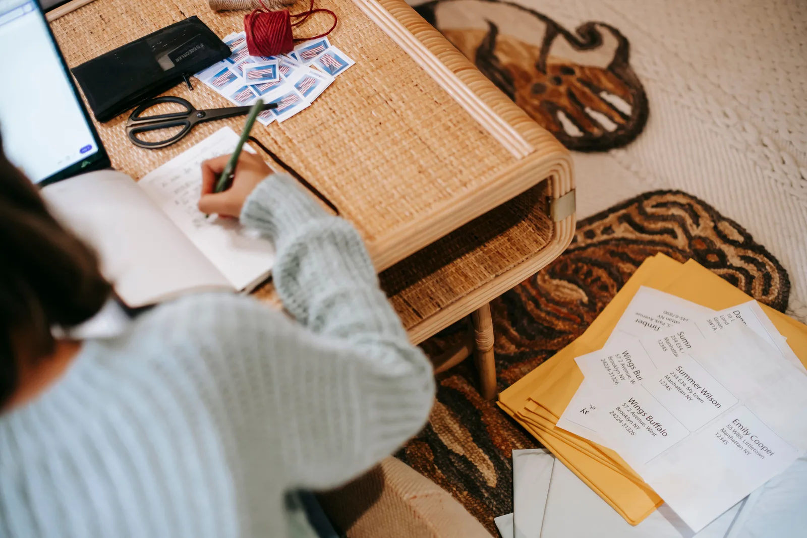High angle of unrecognizable ethnic woman in casual outfit taking notes in notepad at table near laptop and scissors with stamps and shipping parcel bags on floor in light apartment. Money questions worth asking before you put down a creative retreat deposit
